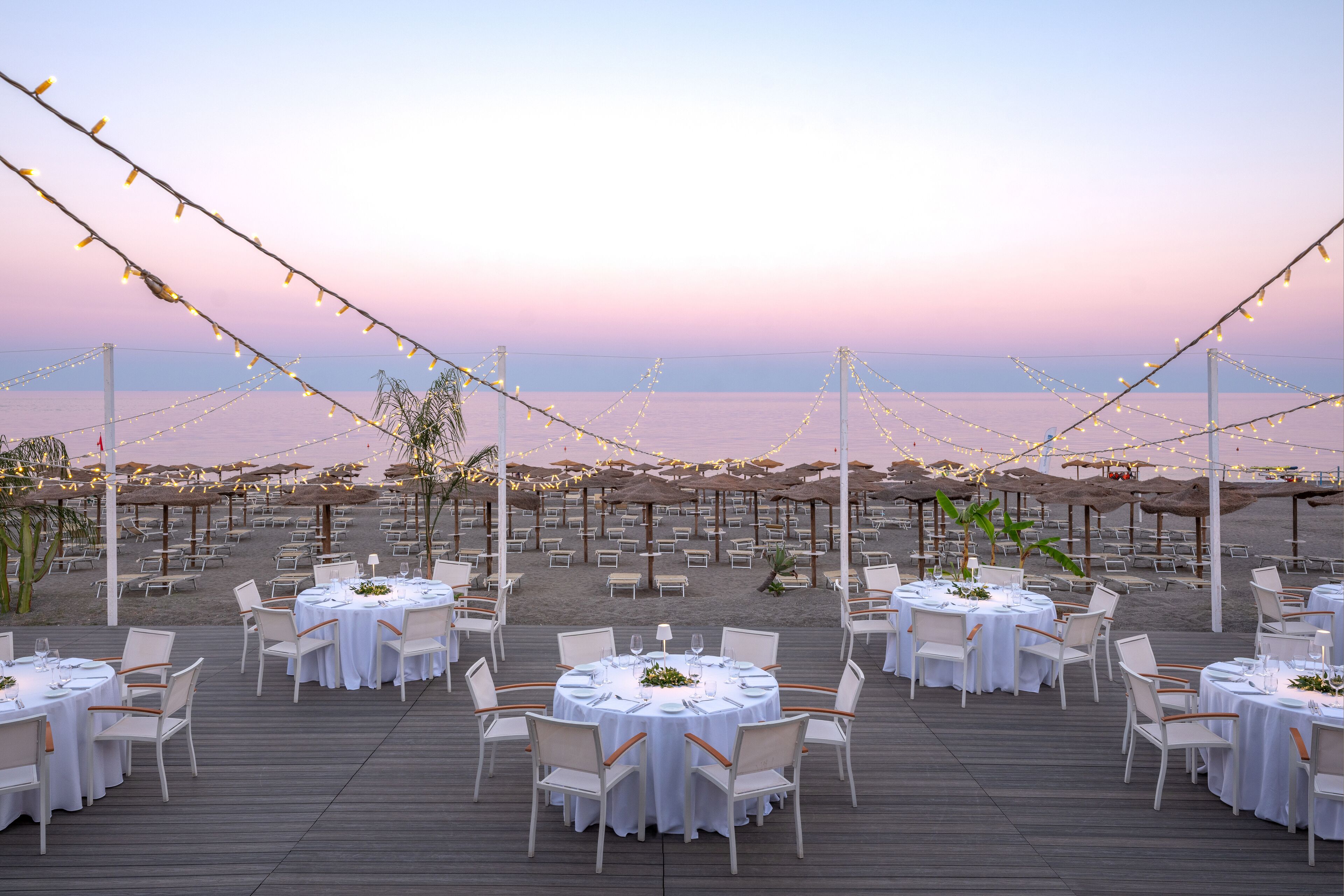 An outdoor dining area with round tables and chairs, string lights, and umbrellas, overlooking a beach.