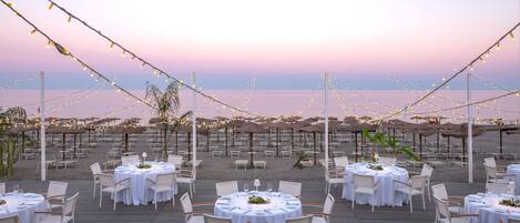 An outdoor dining area with round tables and chairs, string lights, and umbrellas, overlooking a beach.