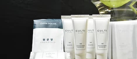 Presentación de artículos de aseo de hotel, incluyendo una bolsa etiquetada 'CUFFIA DOCCIA' y varios tubos de productos 'CULTI MILANO' como champú y gel de ducha.