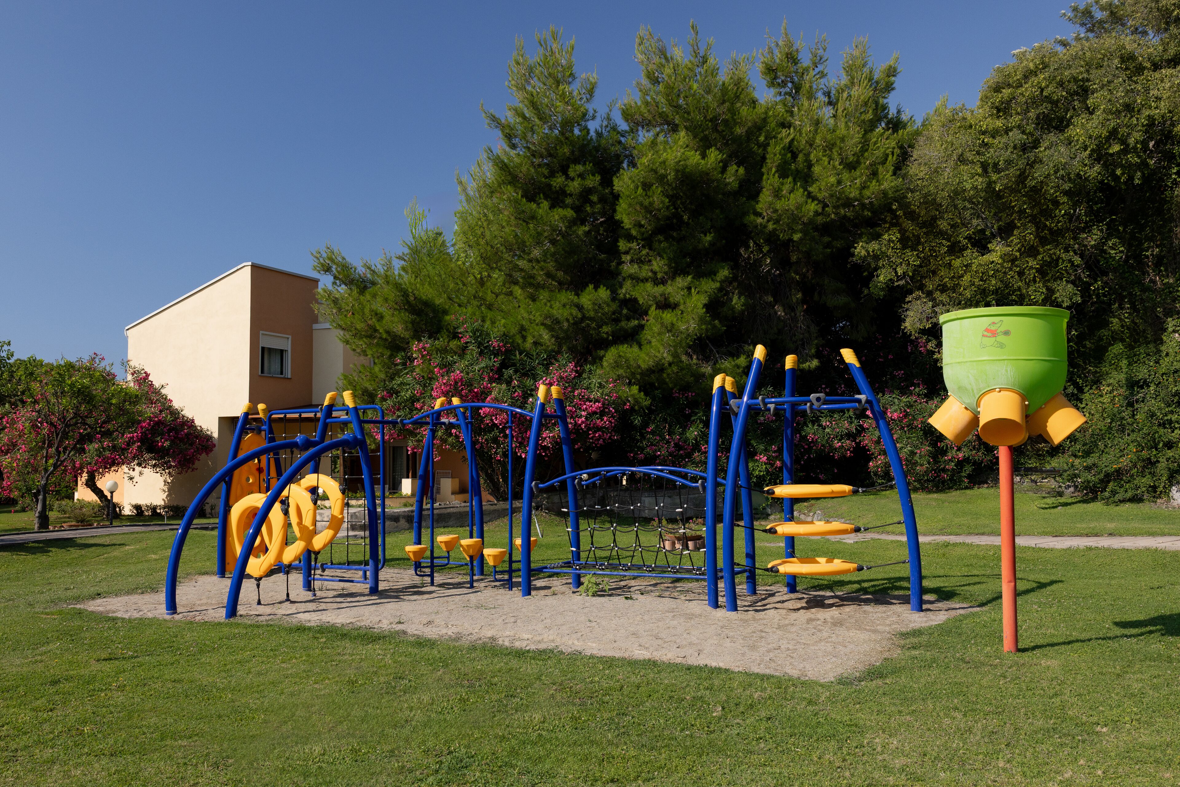 A playground with climbing structures and a sandpit, surrounded by trees and a building.