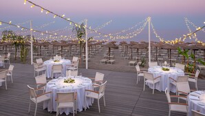 An outdoor dining area with round tables and chairs, string lights, and umbrellas on a wooden deck.