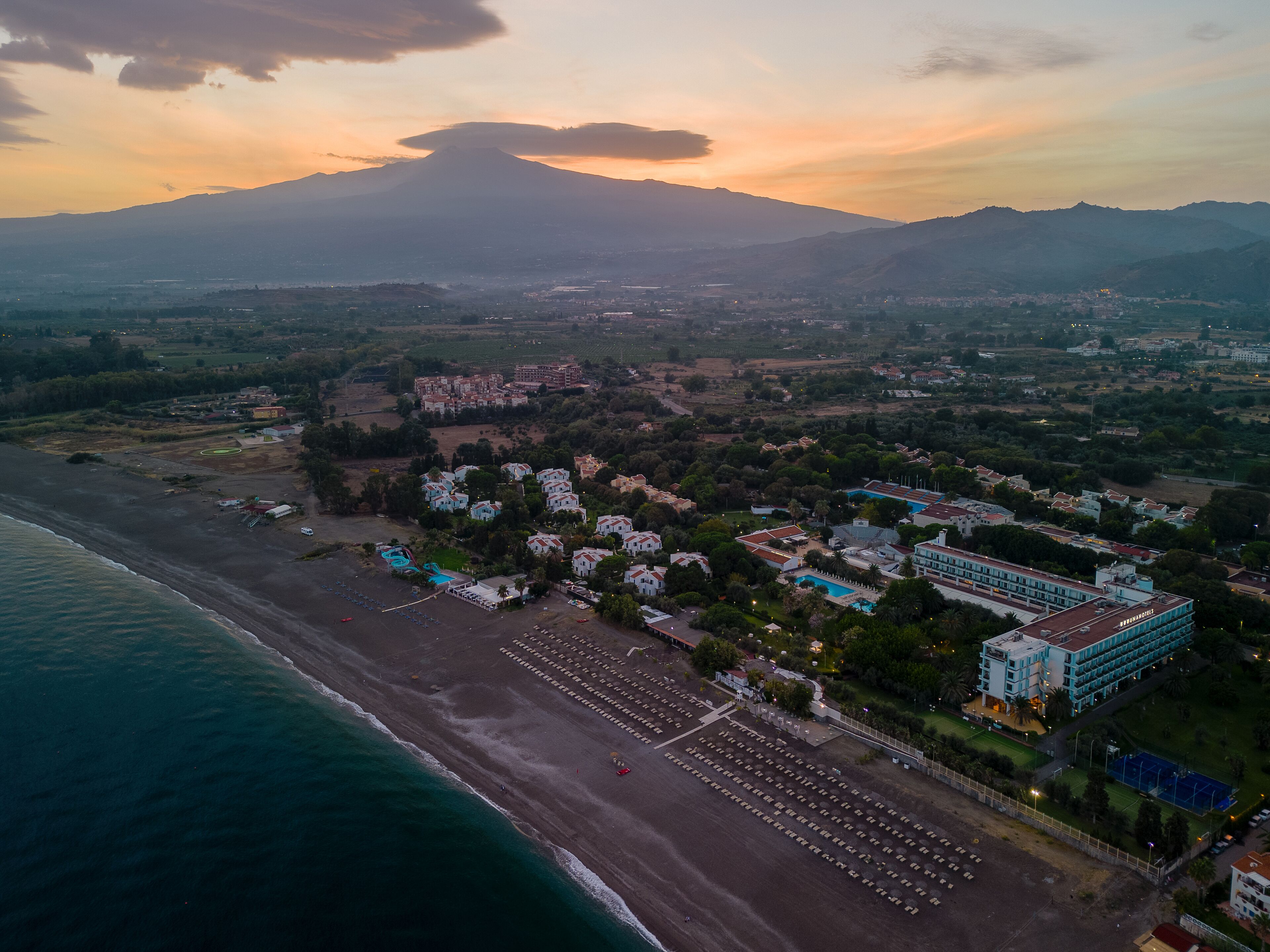 Vista aérea de uma área costeira com um grande complexo hoteleiro, uma praia com fileiras de guarda-sóis e uma montanha ao fundo.