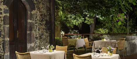 An outdoor dining area with wicker chairs and tables set up against a stone wall, surrounded by greenery.
