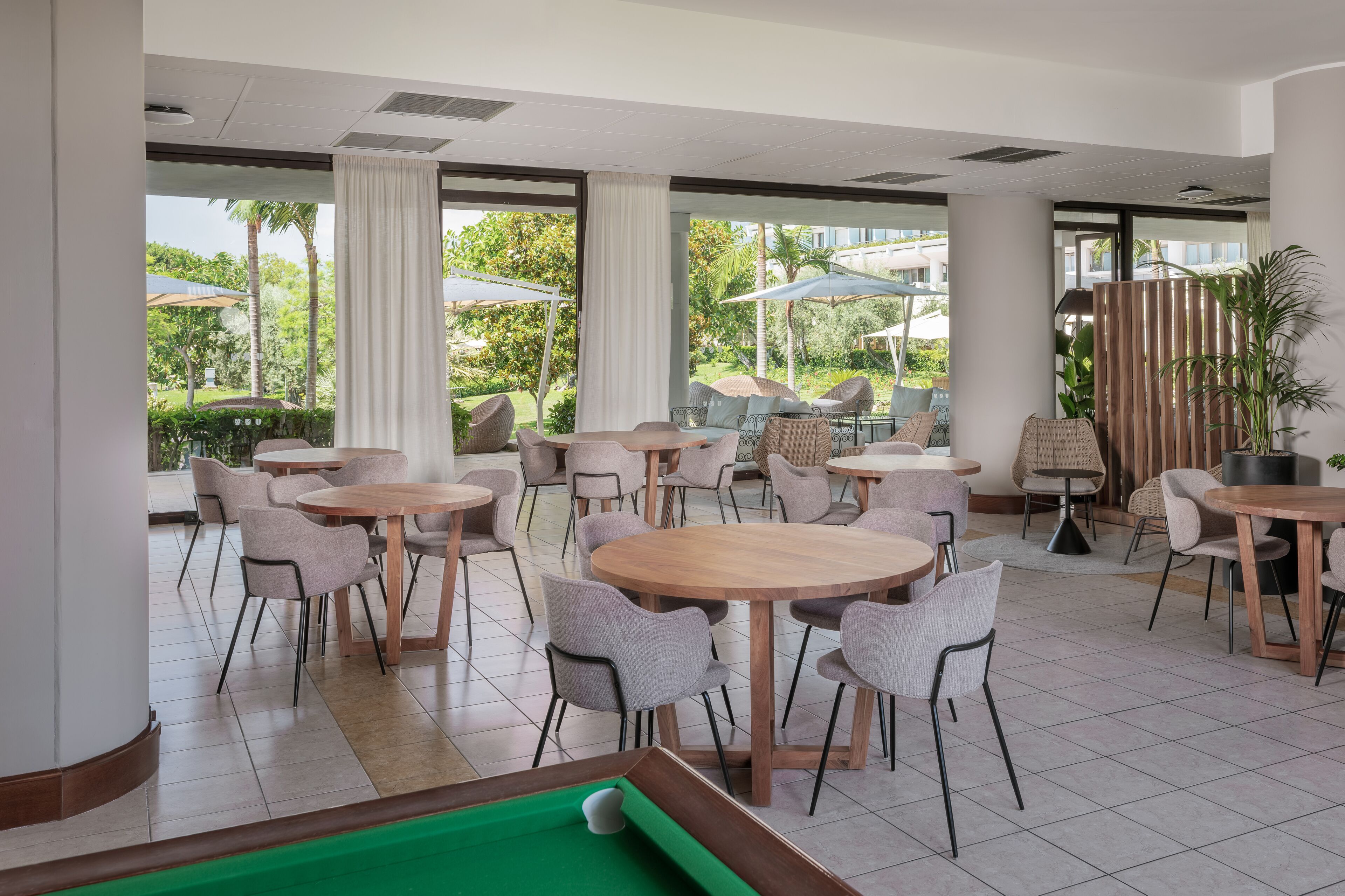A spacious indoor area with multiple round tables and chairs, a pool table, and large windows offering views of greenery and buildings outside.