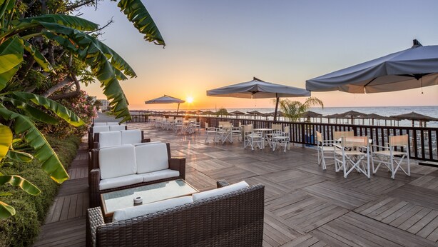 A wooden deck with wicker furniture and umbrellas overlooking the ocean at sunset.