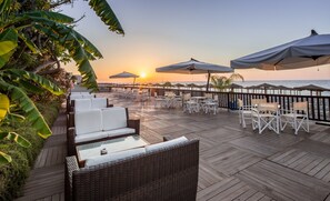 A wooden deck with wicker furniture and umbrellas overlooking the ocean at sunset. - UNA Hotels Naxos Beach Sicilia (Giardini Naxos)