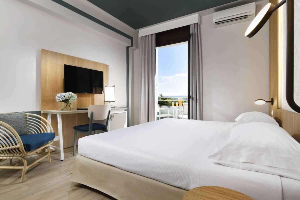 A hotel room with a bed, a desk, a chair, a TV, and a view of the outdoors.