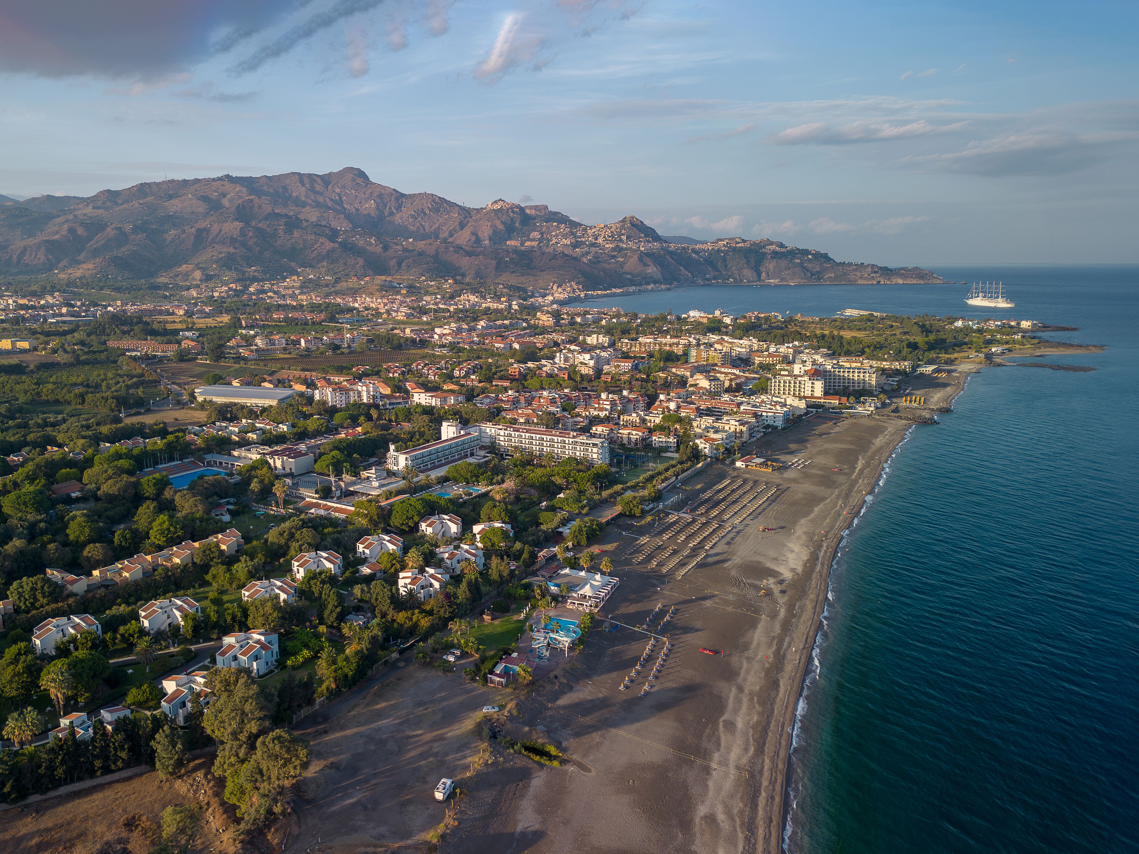 An aerial view of a coastal town with a beach, residential areas, and a mountainous backdrop.