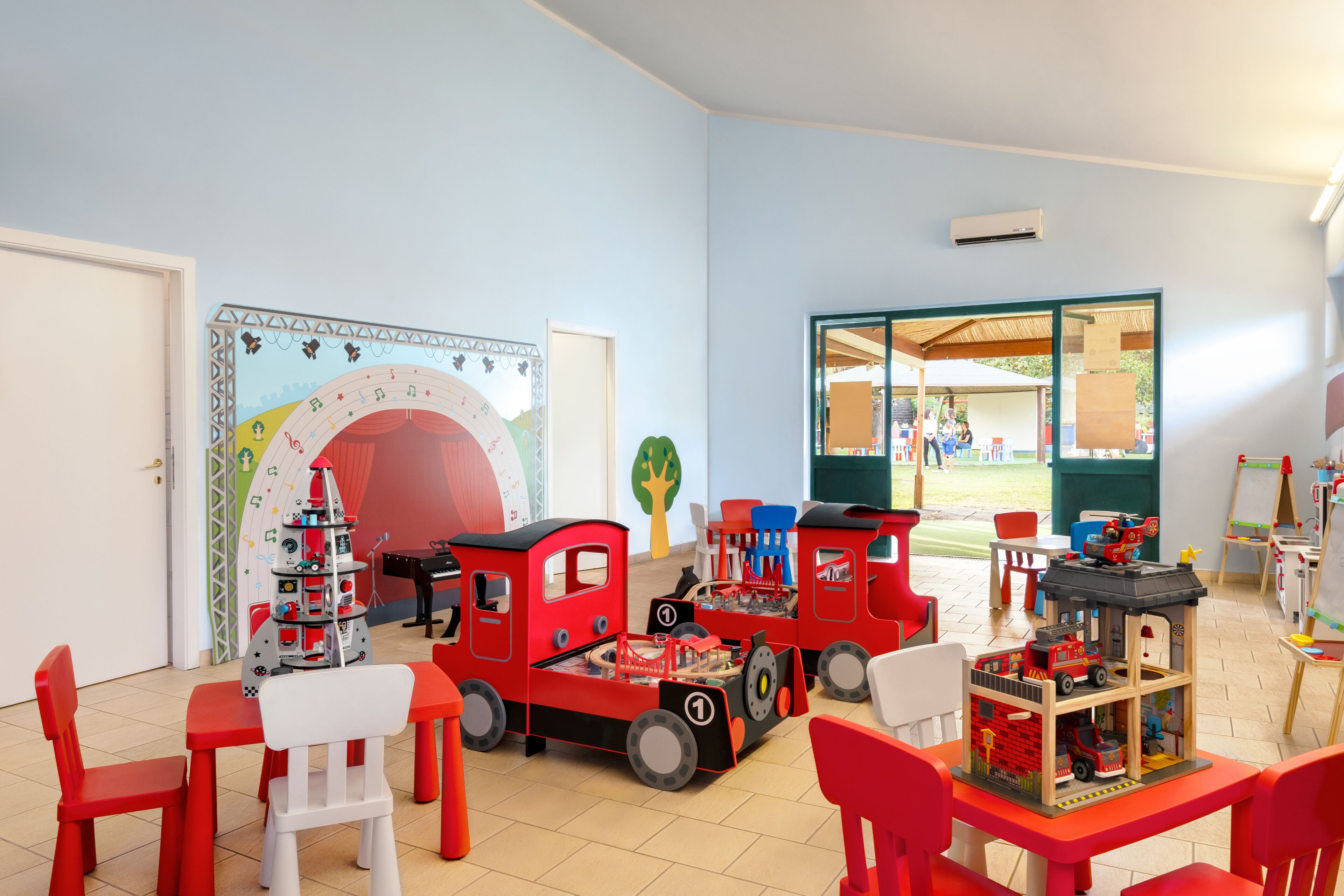A children's playroom with a train set, tables, chairs, and a mural on the wall.