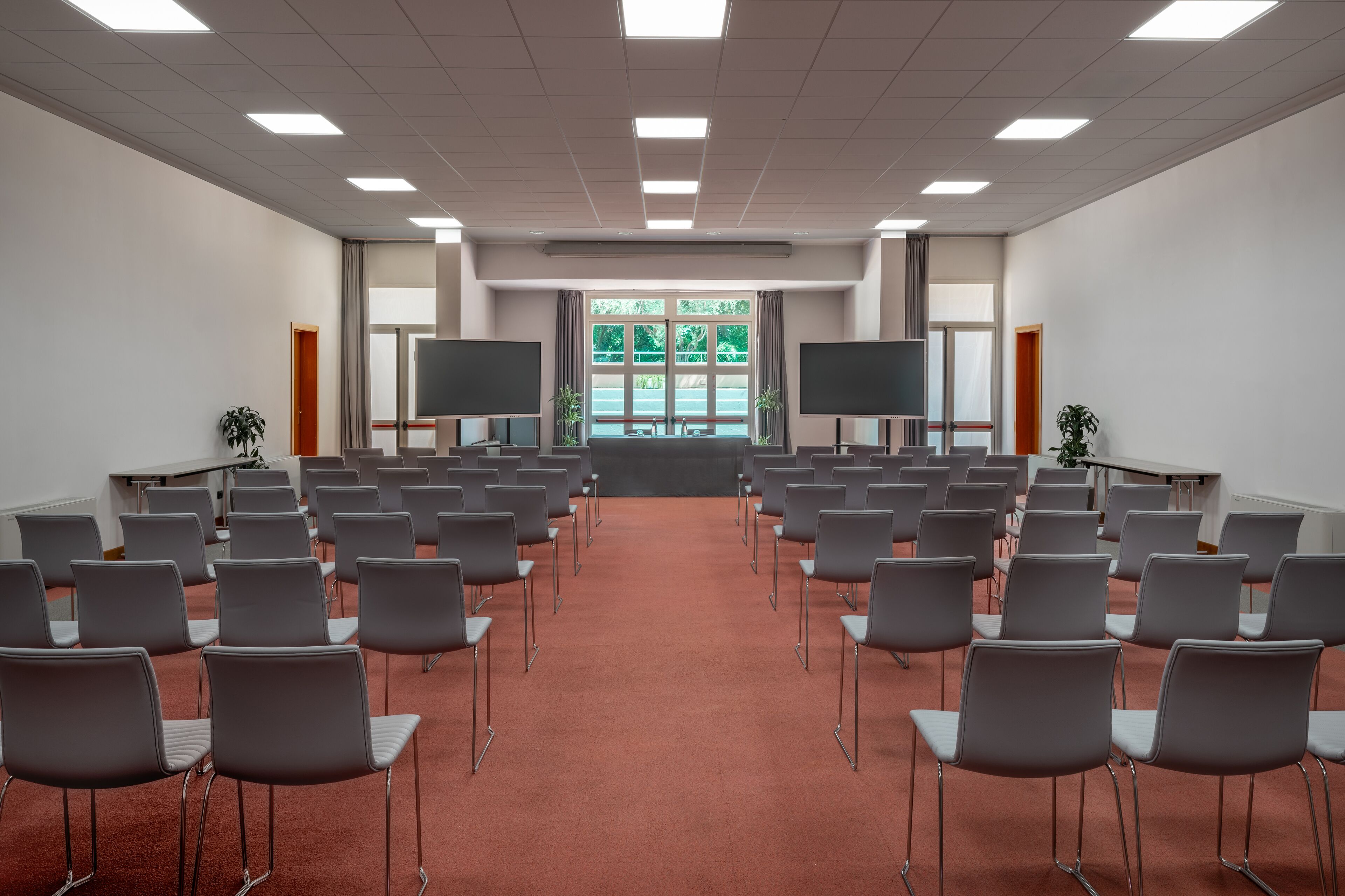 Un salón de conferencias con filas de sillas grises, una alfombra roja, dos pantallas grandes y una ventana con follaje verde.