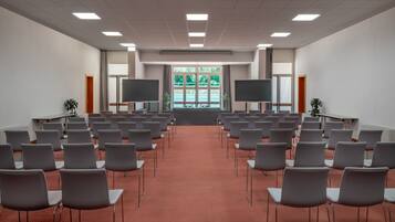 Un salón de conferencias con filas de sillas grises, una alfombra roja, dos pantallas grandes y una ventana con follaje verde.
