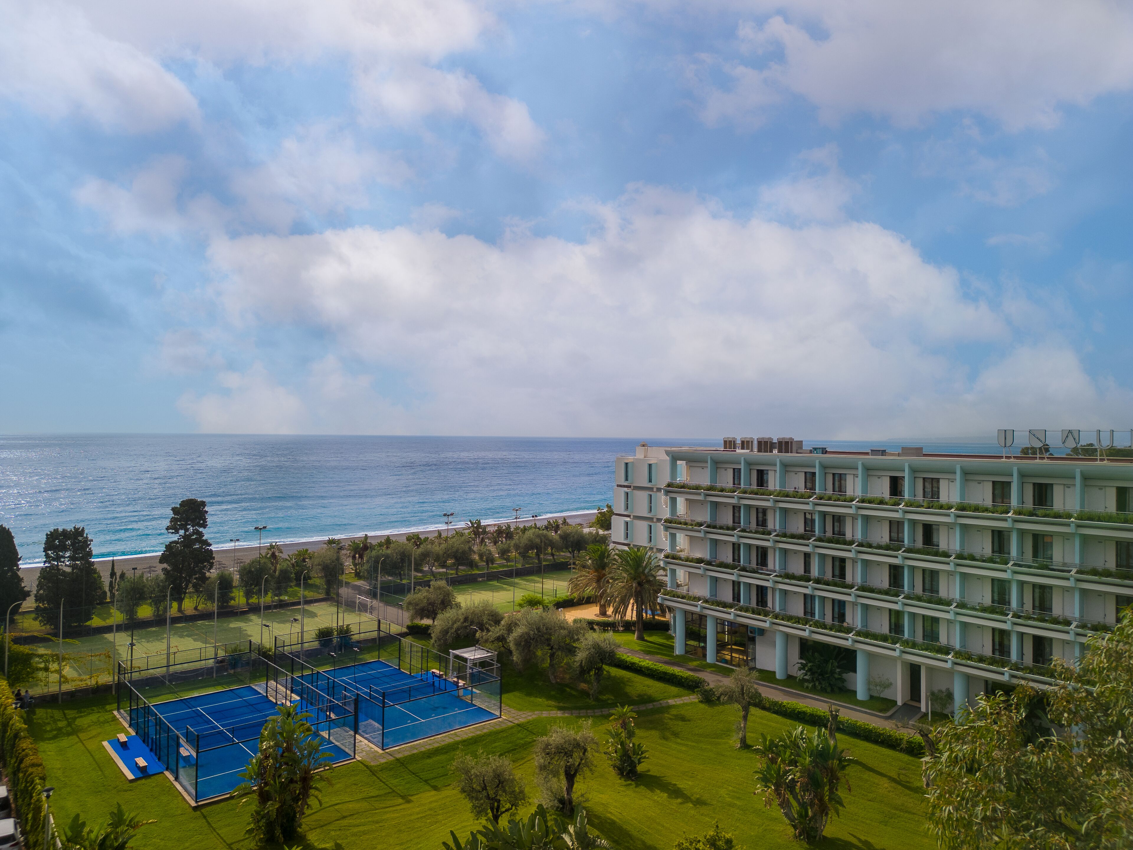 A modern building with a pool and tennis court, overlooking a beach and ocean.