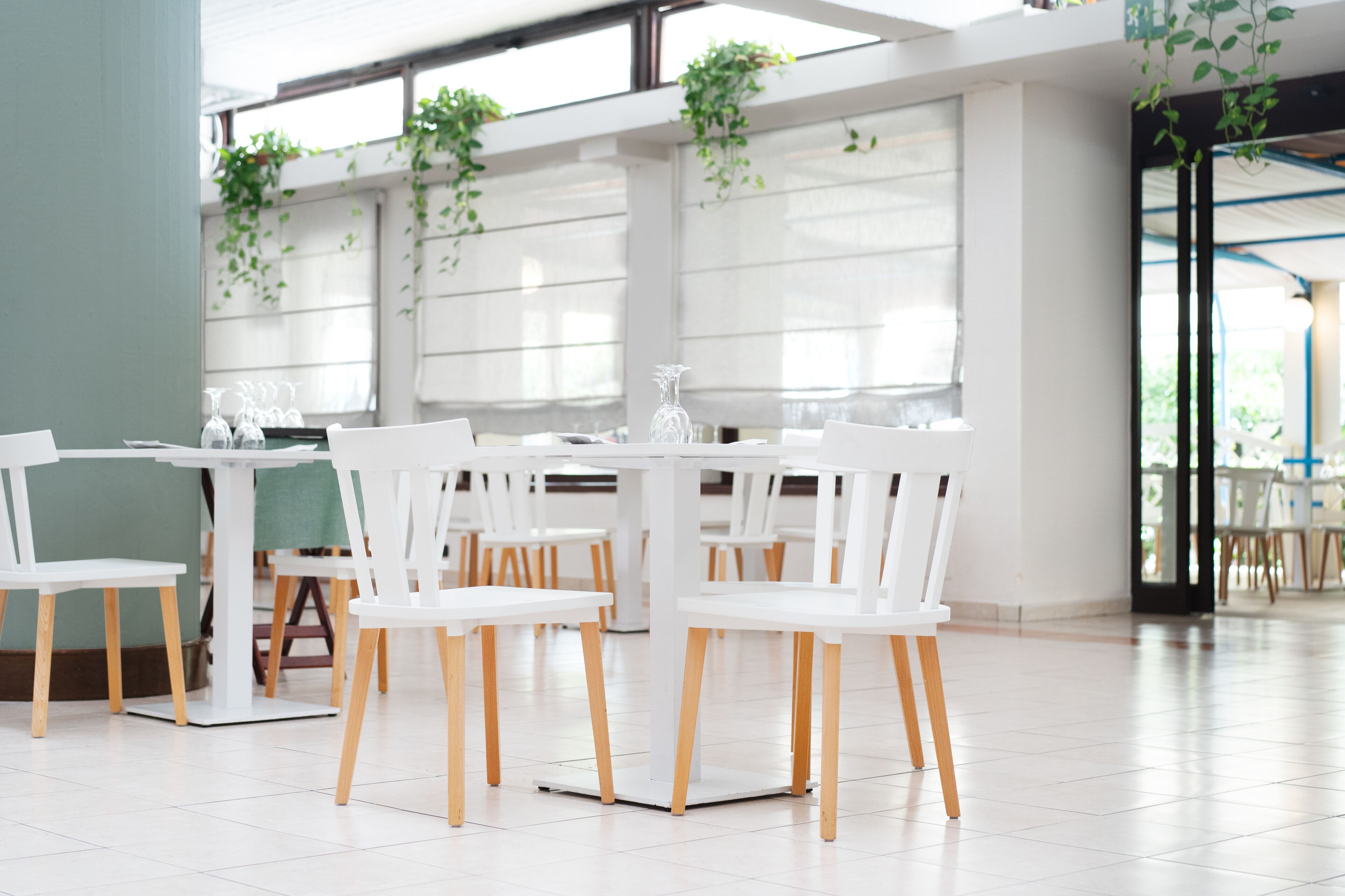 A modern dining area with white tables and chairs, large windows, and hanging plants.