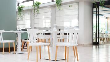 A modern dining area with white tables and chairs, large windows, and hanging plants.