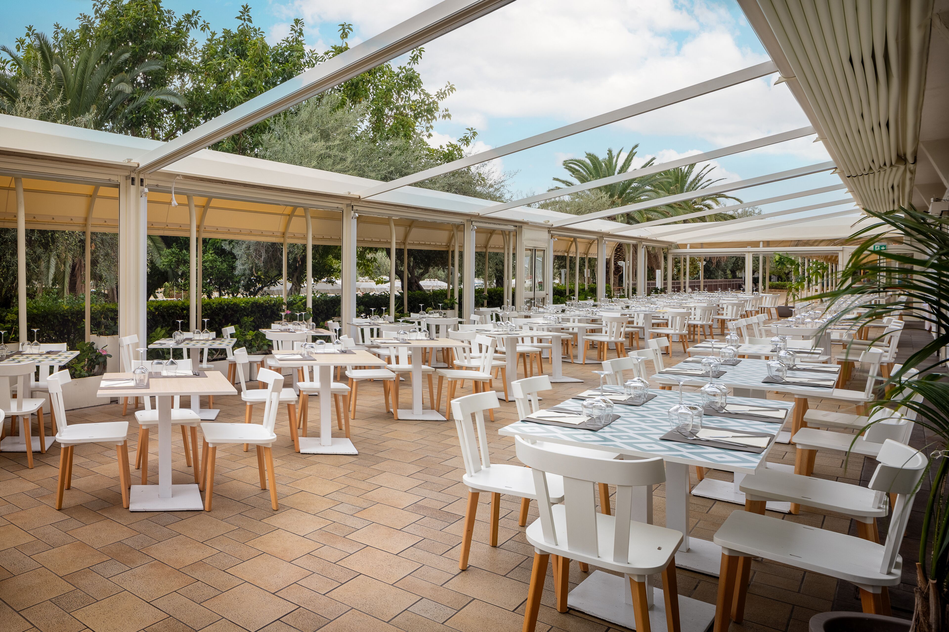 A spacious outdoor dining area with white tables and chairs, surrounded by glass panels and greenery.