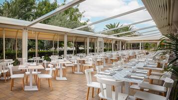 A spacious outdoor dining area with white tables and chairs, surrounded by glass panels and greenery.