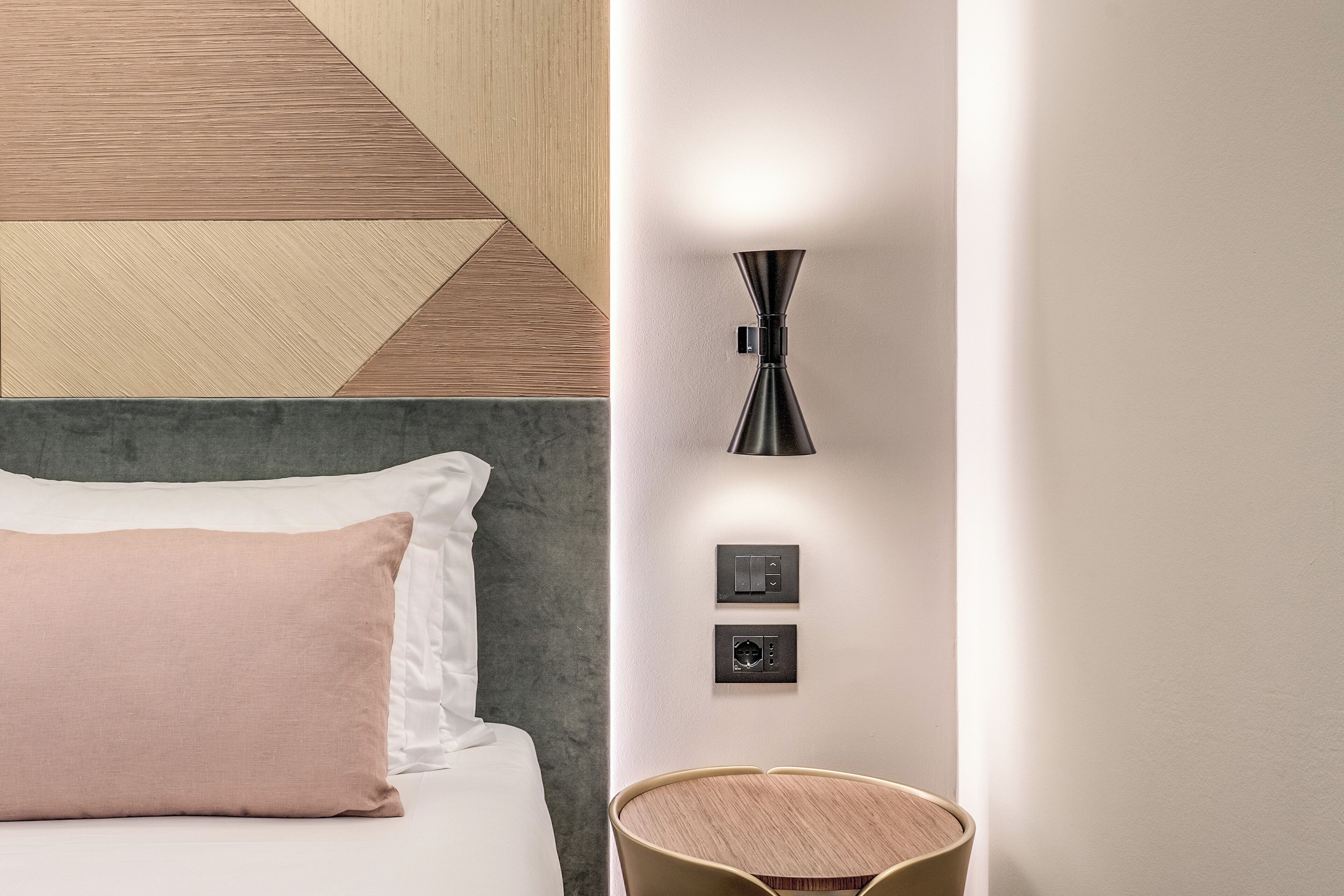 A modern hotel room with a bed, a wall-mounted light fixture, and a small wooden side table.
