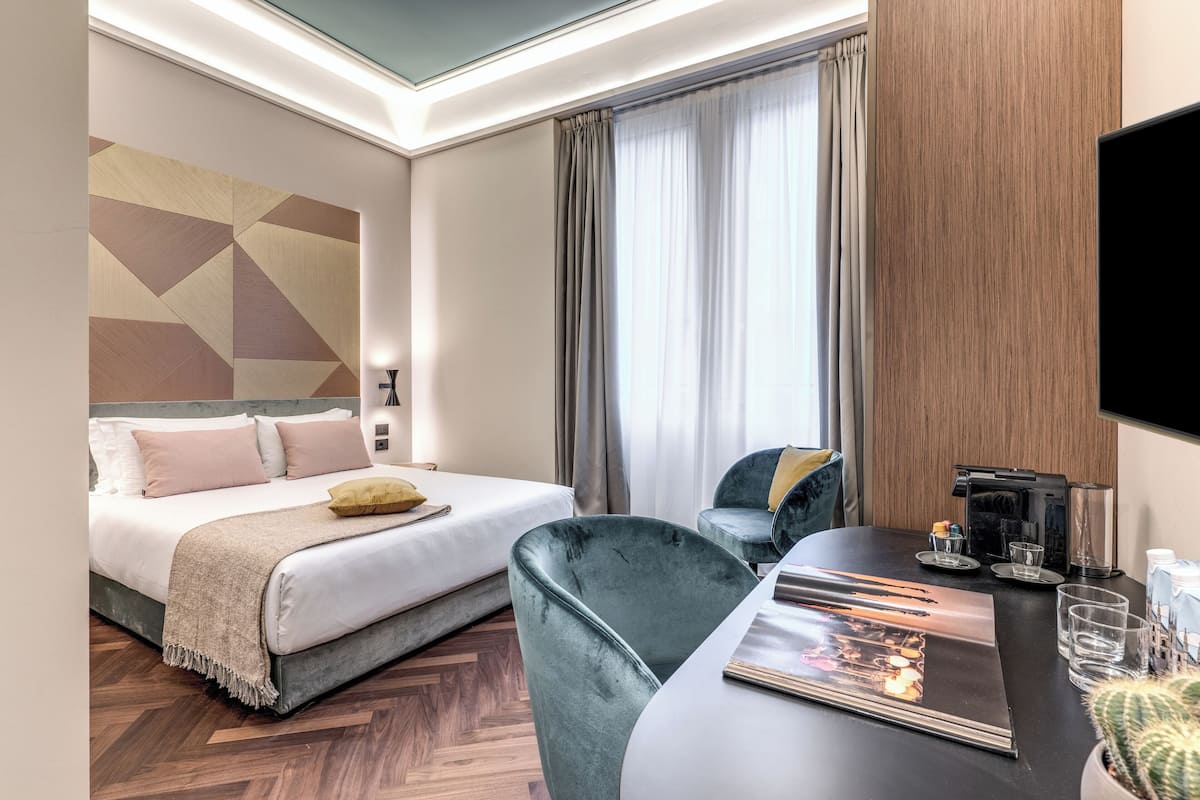 Habitació d'hotel amb un llit, un escriptori, una cadira, un televisor, una cafetera i un cactus.