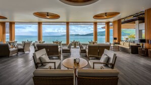 Amplia sala de estar con vista al mar, con mobiliario de mimbre, una mesa redonda central y un techo con paneles de madera y luces colgantes.
