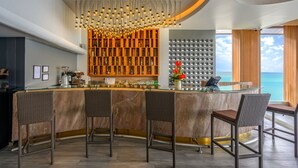 Zona de bar con una barra de mármol curvada, taburetes de mimbre, una lámpara moderna y una pared de unidades de almacenamiento decorativas.