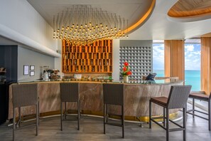 A bar area with a curved marble countertop, wicker bar stools, a modern chandelier, and a wall of decorative storage units. - Namaka Resort Kamala (Kamala)