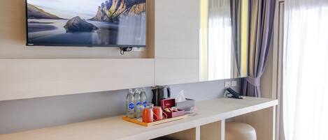 A modern hotel room with a flat-screen TV mounted on the wall, a wooden desk with a tray of amenities, a minibar, and a small black refrigerator.