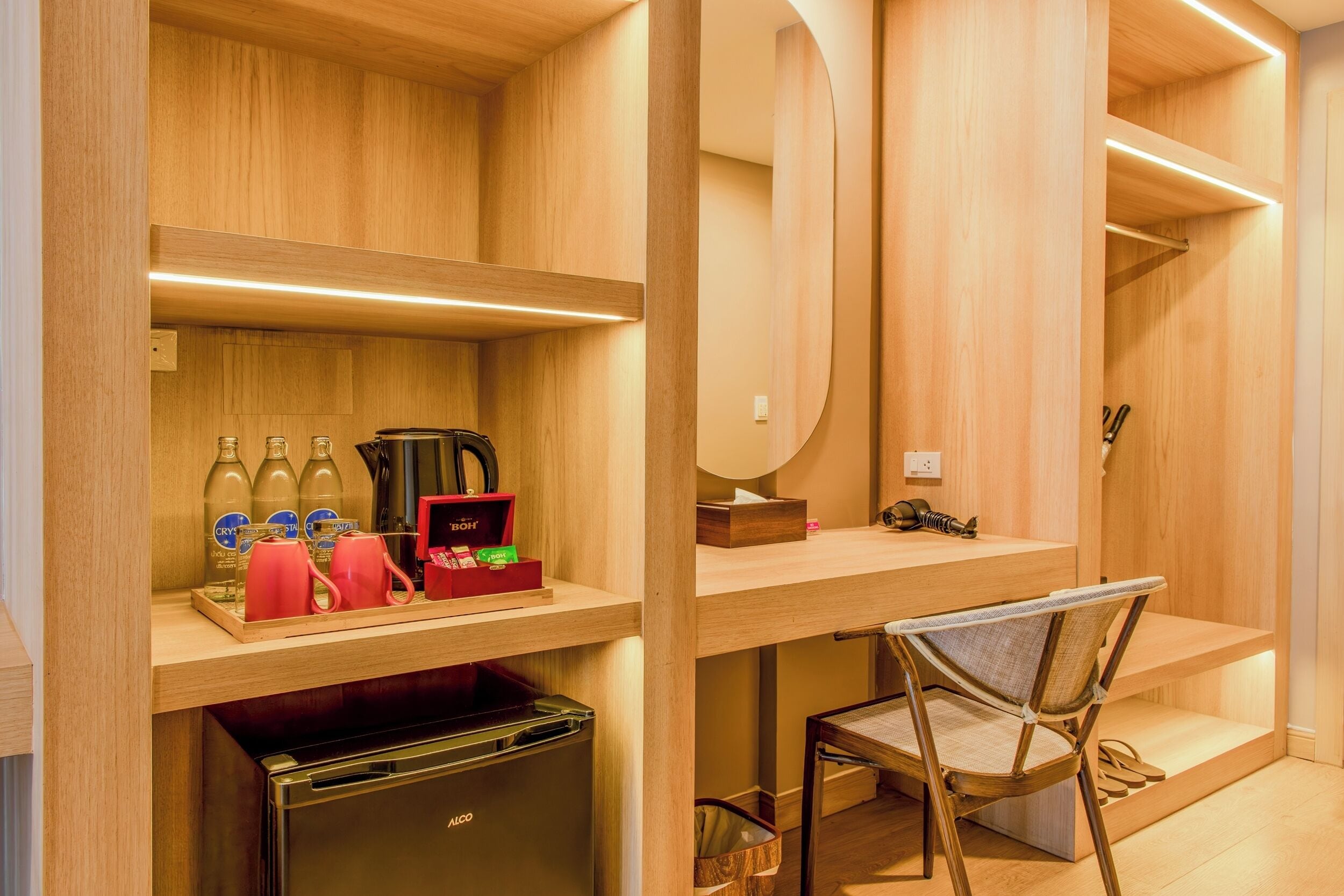 A modern hotel room with wooden paneling, a desk with a chair, a minibar, and a built-in mirror.