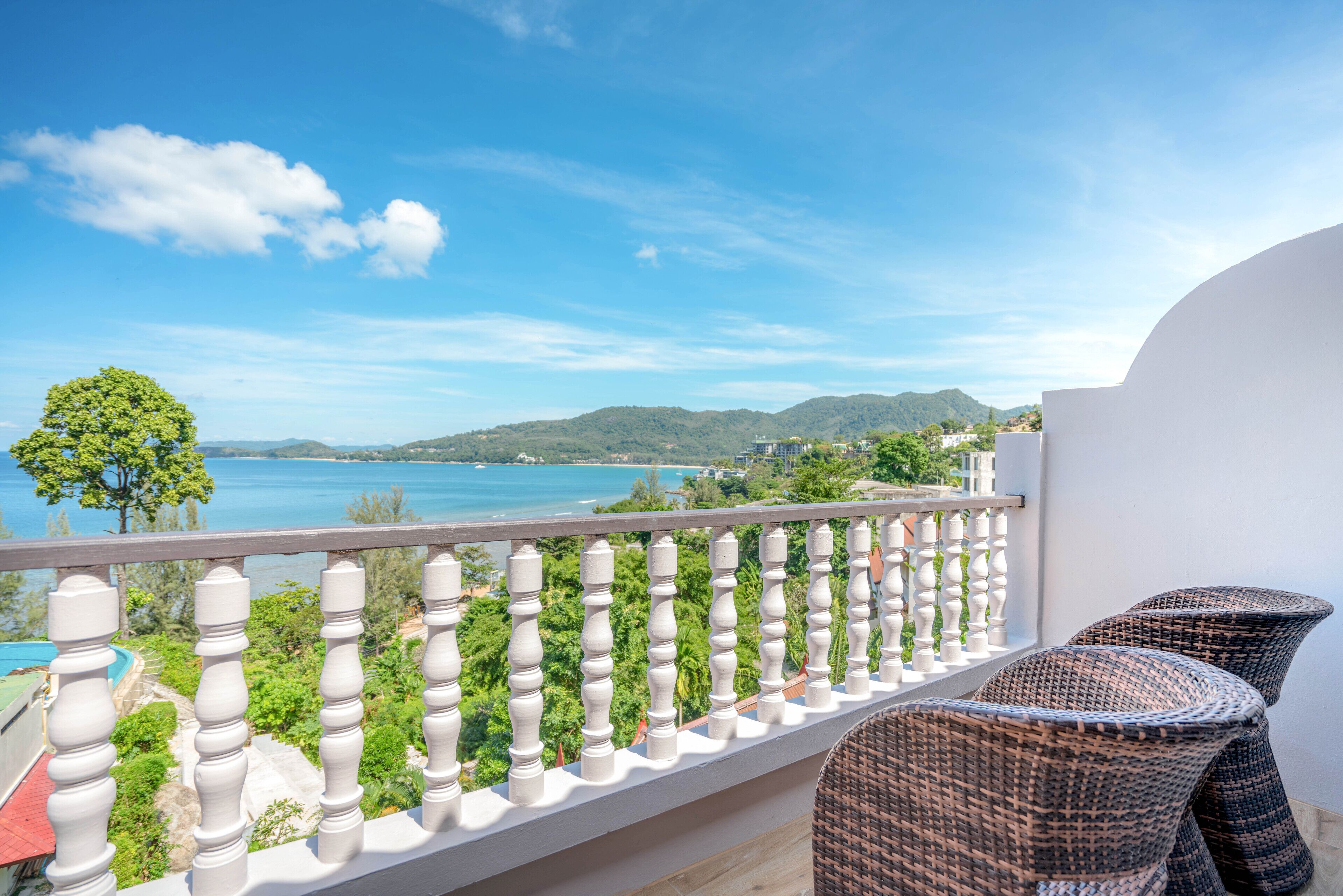 Un balcone con sedie in vimini, una ringhiera bianca e vista su un paesaggio costiero con colline e uno specchio d'acqua.