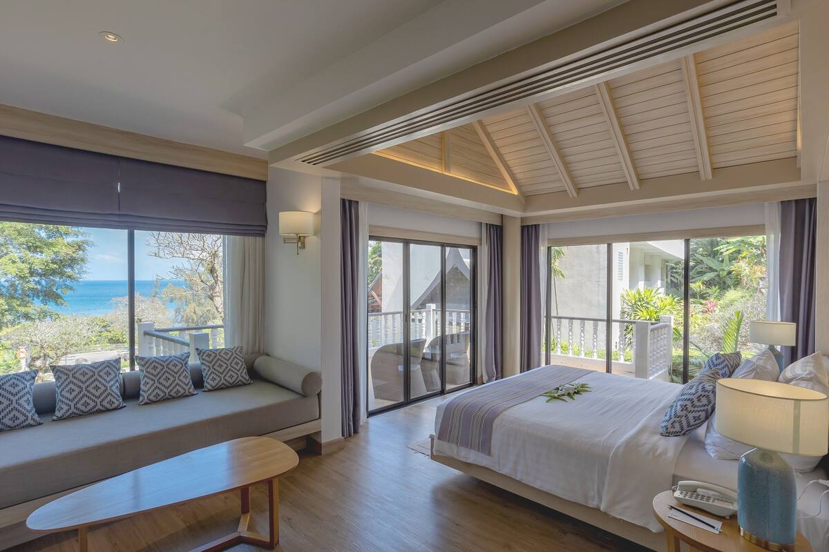 A bedroom with a large bed, a seating area with cushions, a bedside table with a lamp, and a view of the outdoors.