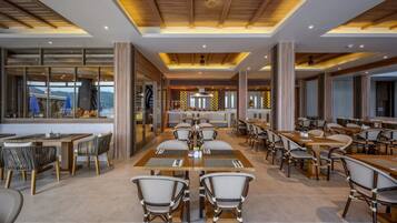A spacious dining area with multiple tables and chairs, large windows, and a modern ceiling design.