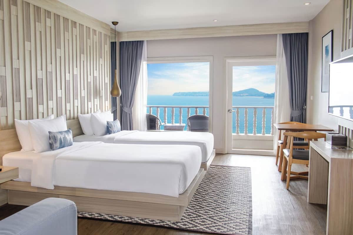 A hotel room with two beds, a desk, and a balcony with a view of the sea.