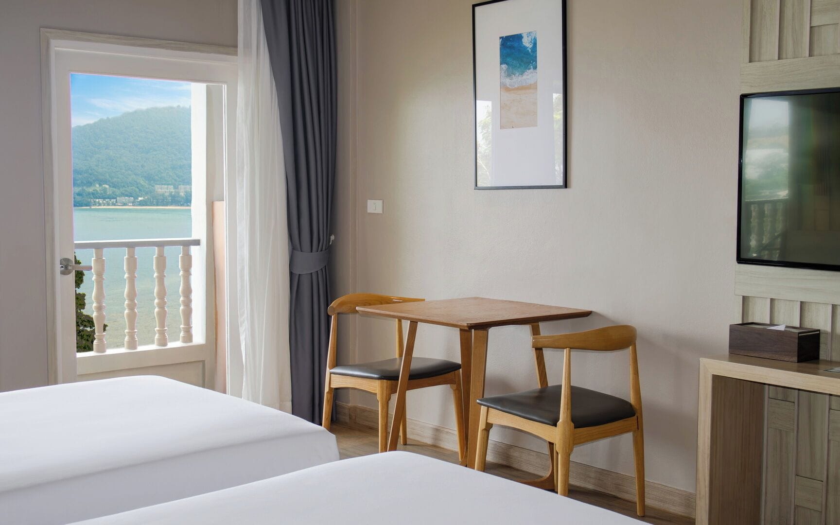 A hotel room with a balcony, a wooden table with two chairs, a bed, a TV, and a framed picture on the wall.