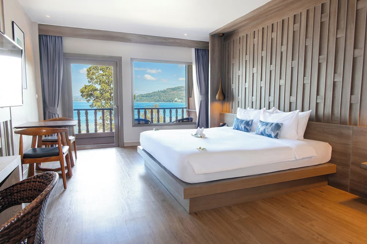 A modern hotel room with a large bed, a dining table with chairs, and a view of the sea through the balcony.