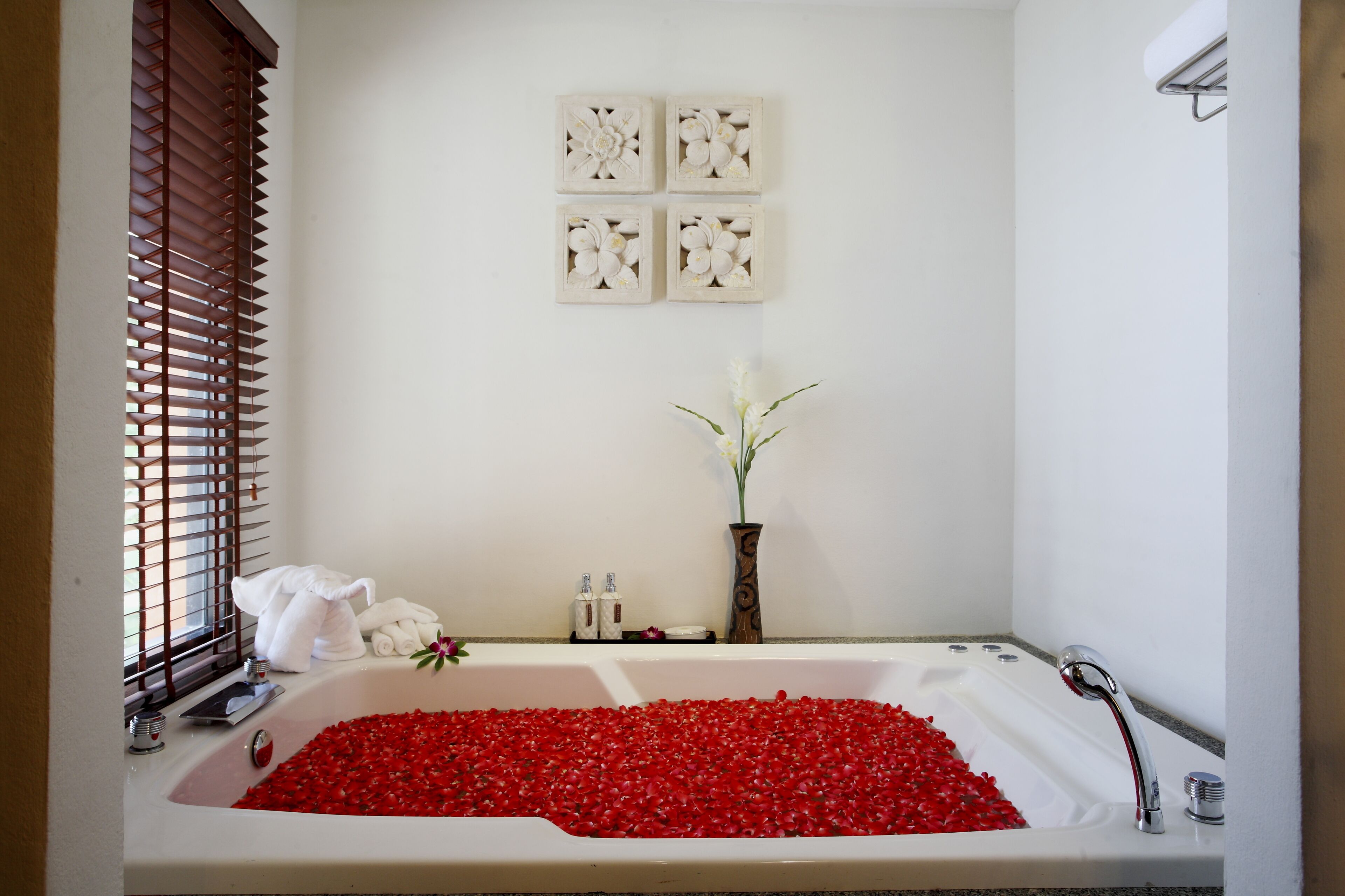 A bathroom with a bathtub filled with red rose petals, a white vase with flowers, and a window with wooden blinds.