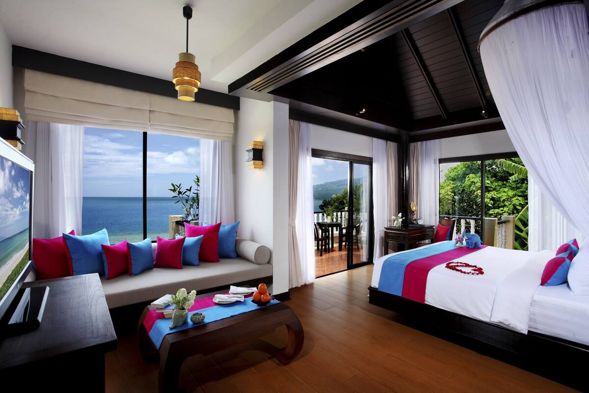 A hotel room with a large window, a sofa with colorful cushions, a bed with a red heart design, and a wooden coffee table.