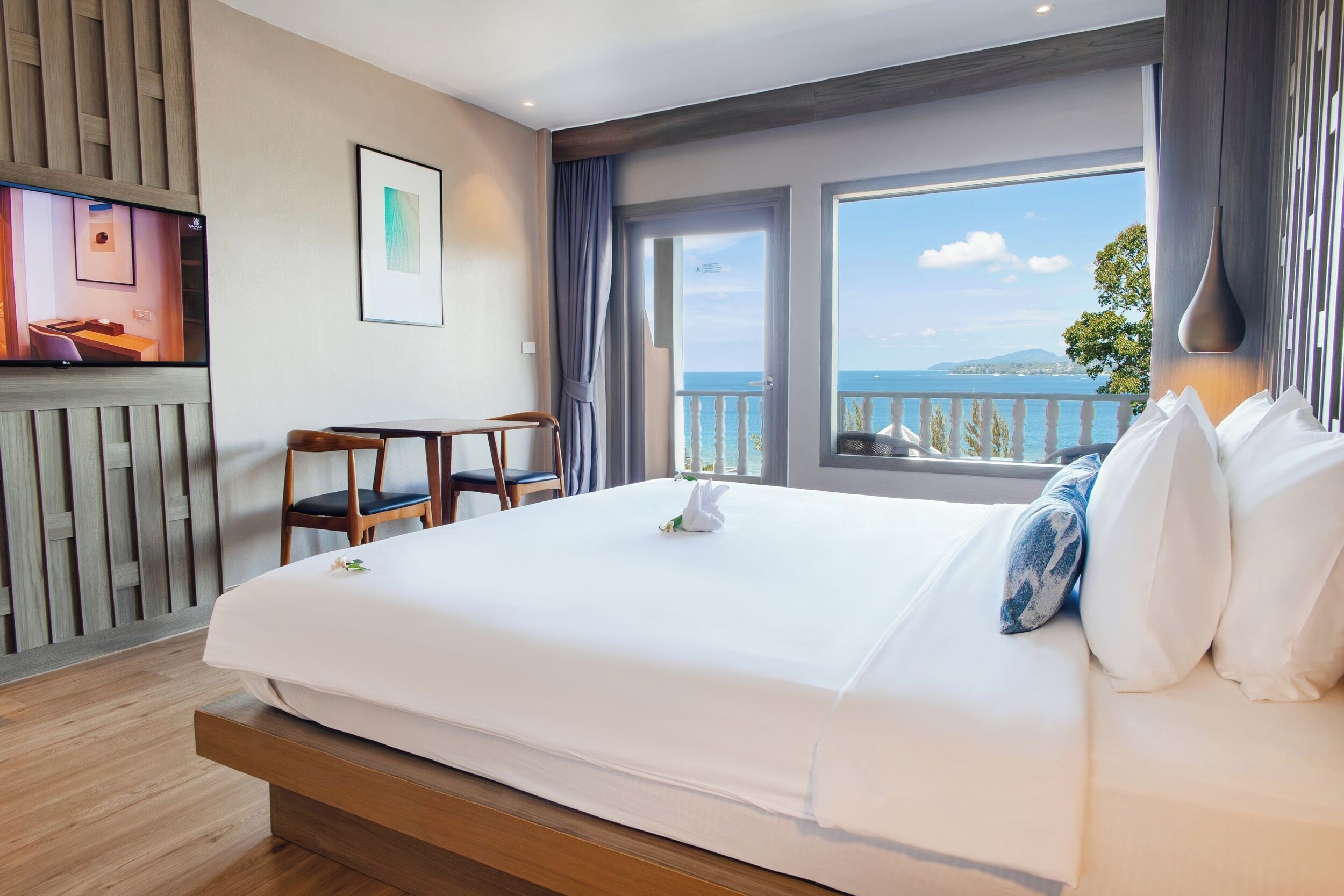 A hotel room with a large bed, a TV on the wall, a desk, and a balcony with a view of the sea.