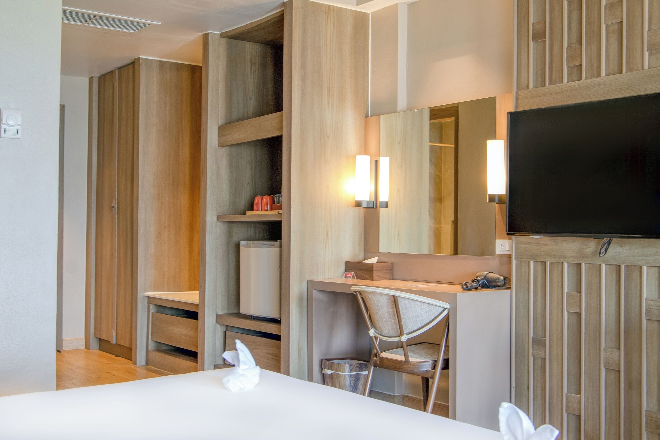 A modern hotel room with a wooden wardrobe, a desk with a chair, a flat-screen TV, and a small trash bin.