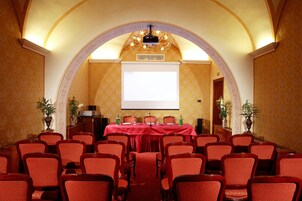 Un salón de conferencias con alfombra roja, sillas de madera, una pantalla de proyector y una mesa con botellas de agua.