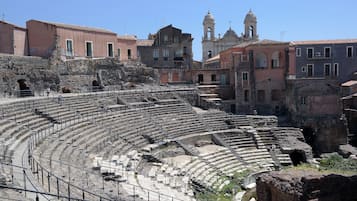 An ancient amphitheater with stone seating and a central stage, surrounded by historic buildings and a bell tower.
