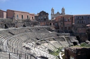 Un antico anfiteatro con gradinate in pietra e un palco centrale, circondato da edifici storici e un campanile.