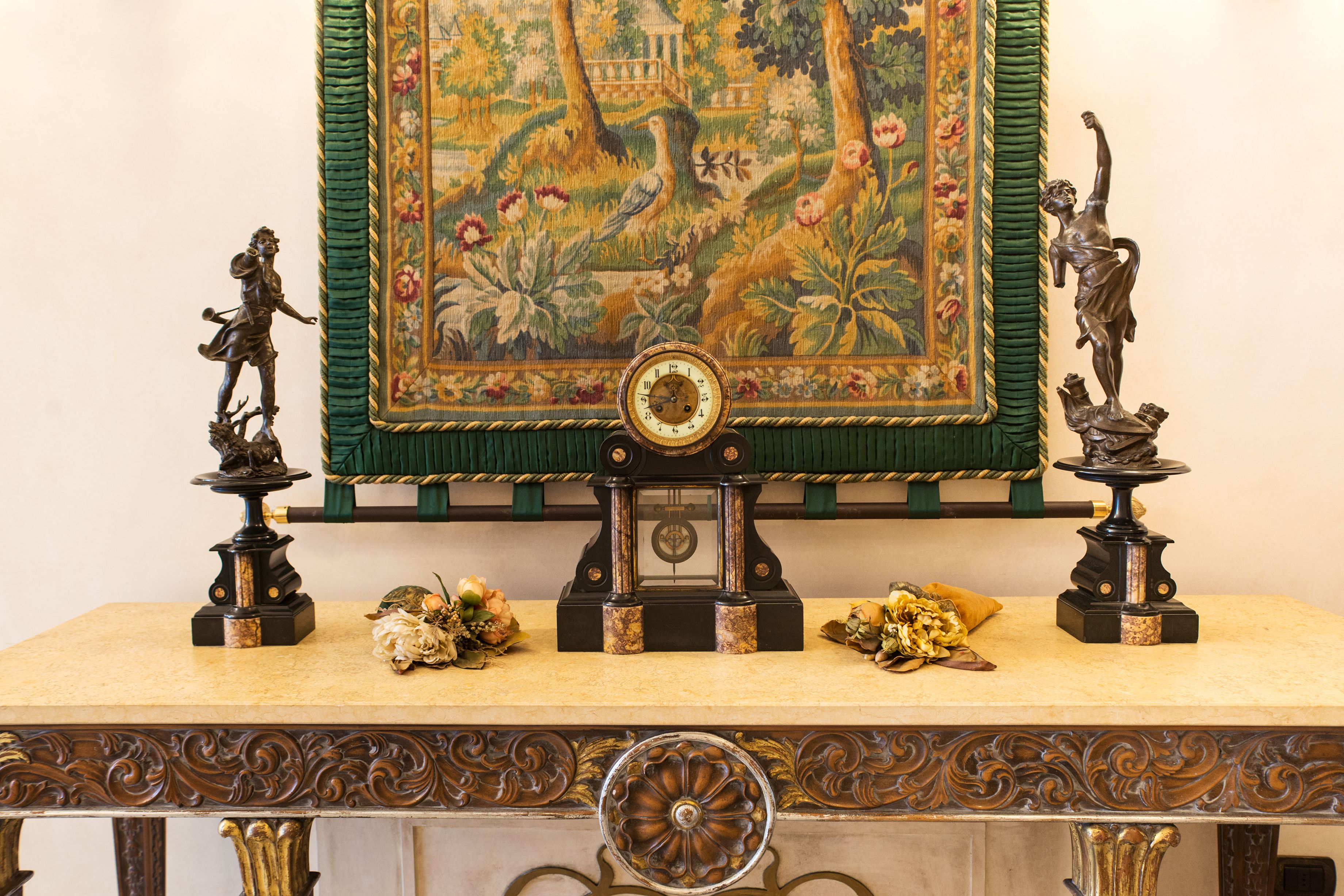 A table with a clock, two statues, and a tapestry.