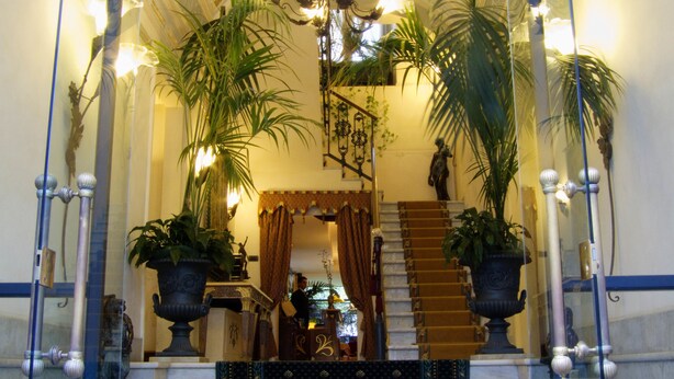 A grand staircase with ornate railings and potted plants, leading to a room with a chandelier and curtains. - Liberty Hotel (Catania)