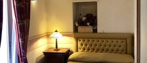 A hotel room with a bed, a nightstand with a lamp, a window with curtains, and a wall-mounted shelf with a vase.