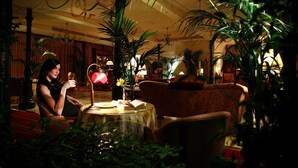 A woman sitting at a table in a warmly lit room with a patterned sofa and several potted plants. - Liberty Hotel (Catania)