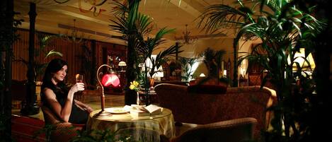 A woman sitting at a table in a warmly lit room with a patterned sofa and several potted plants.
