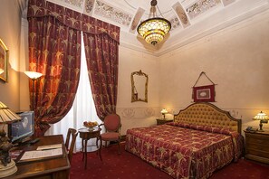 Classic design with a large bed, sitting area, and desk, featuring a chandelier and framed artwork.