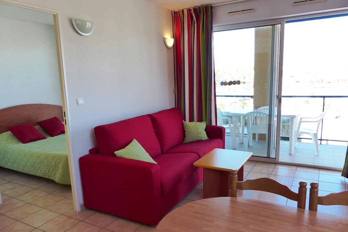 Ein Zimmer mit einem roten Sofa, einem grünen Bett, einem Holztisch und einem Balkon mit weißen Stühlen.