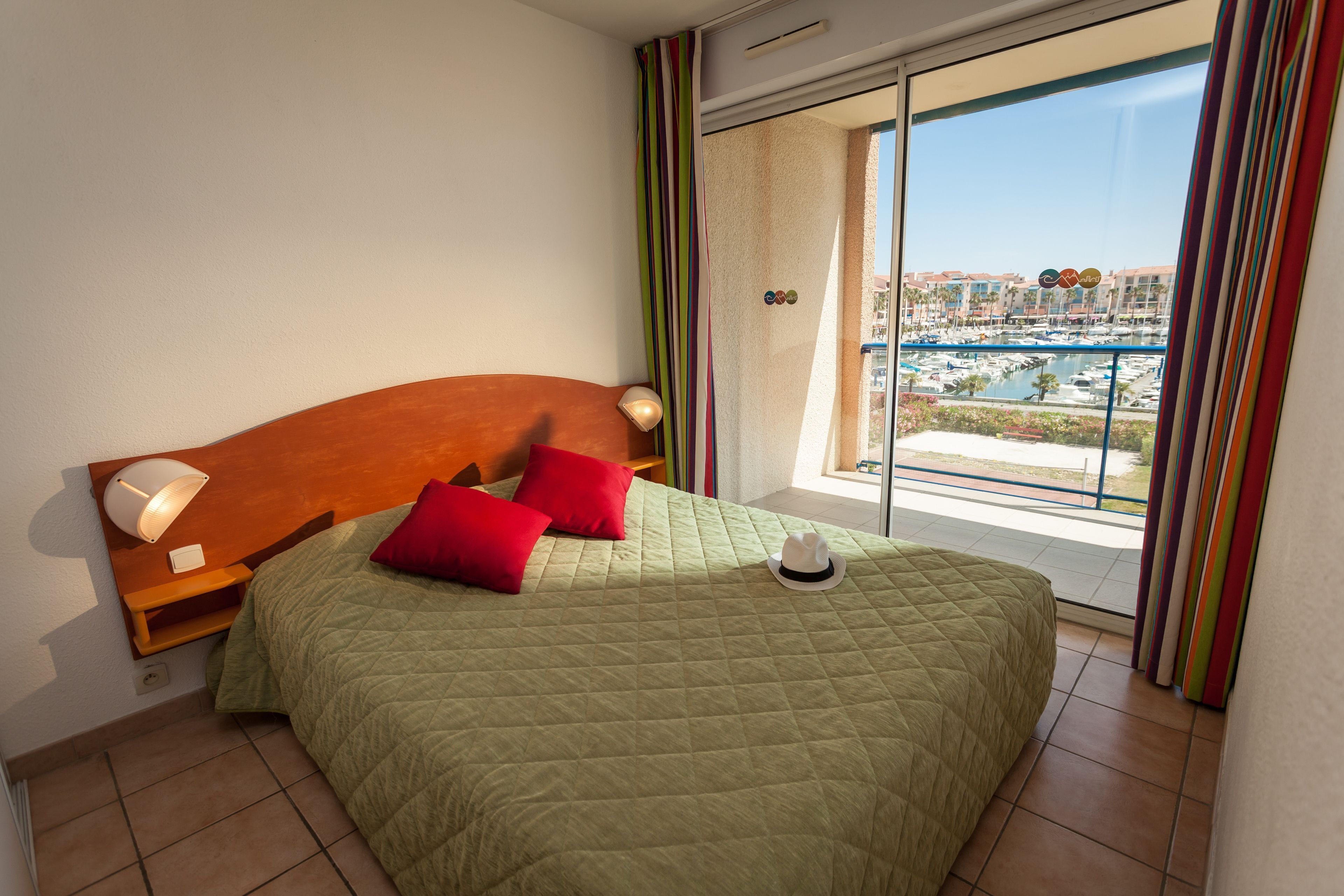 A hotel room with a bed, a nightstand, a lamp, a window with a view of a marina and buildings.