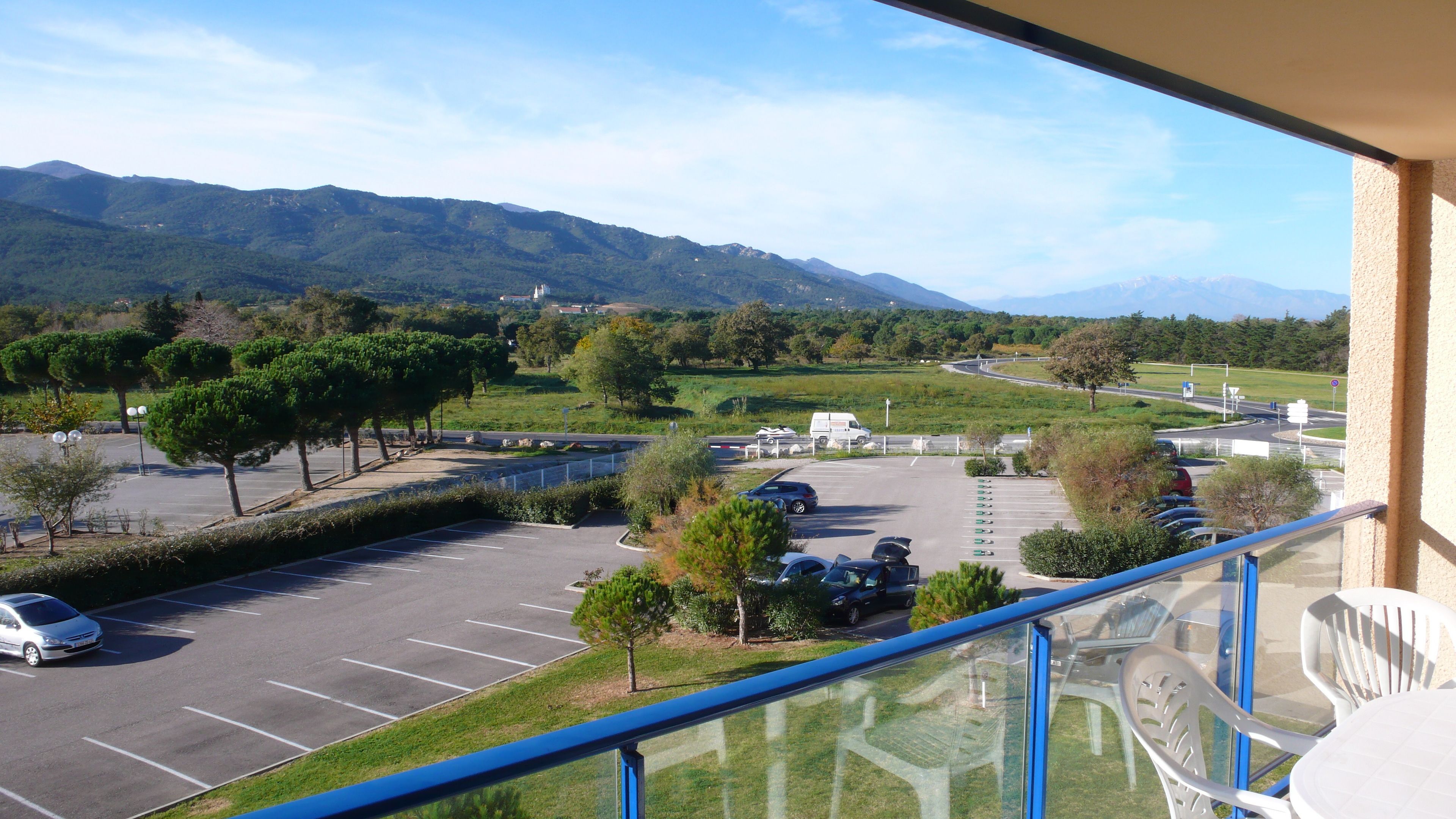 Un balcon donnant sur un parking, des arbres et des montagnes au loin.