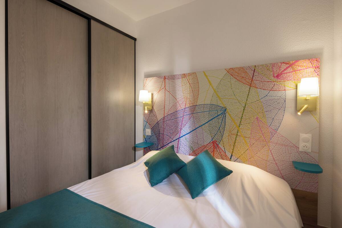 Ein Hotelzimmer mit Bett, zwei Kissen, einem Nachttisch, einer Lampe und einer Wand mit einem farbenfrohen abstrakten Design.